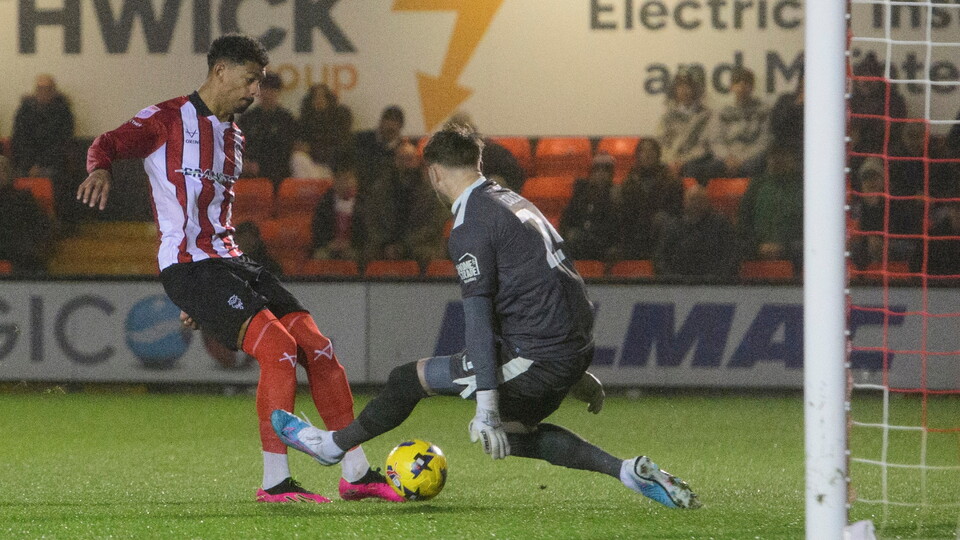 A football player in red and white kit prepares to strike the ball past a goalkeeper in grey.