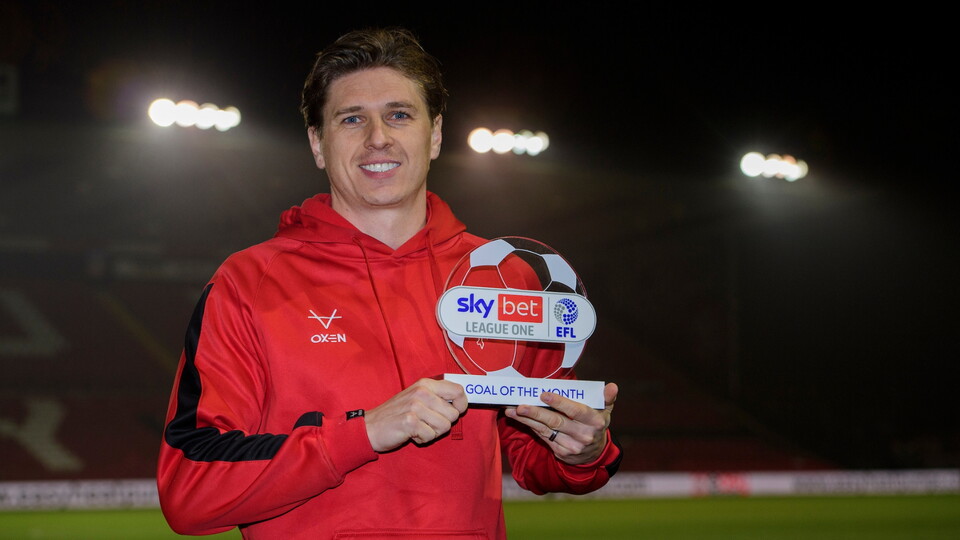 A man in a red hoodie smiling and holding a Sky Bet League One Goal of the Month trophy. Stadium lights are visible in the dark background.