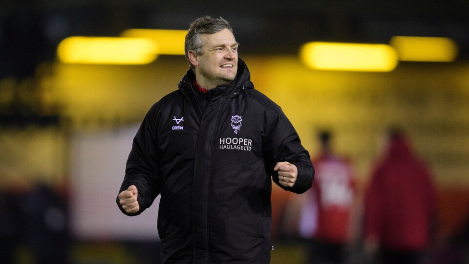 A man in a black jacket smiles confidently, clenching his fists, with a blurred stadium background.