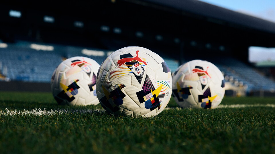 Three footballs with a rainbow motif on them sit on a grass pitch