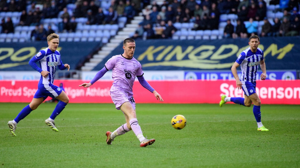 A football player in a violet kit prepares to kick a ball on the field, with two opposing players in blue and white kits approaching. The stadium seats are filled with spectators. 