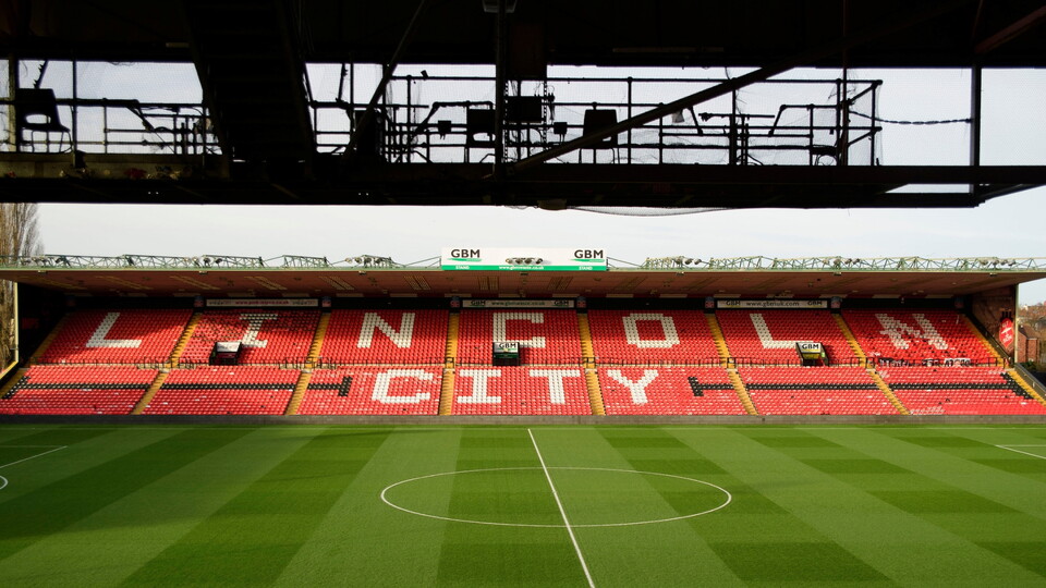 A football stadium with an empty green pitch and red stands displaying "Lincoln City" in bold white letters