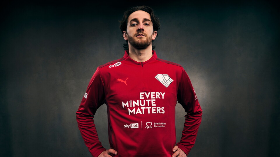 A confident man poses in a red jersey with the slogan Every Minute Matters. The shirt features logos including Puma and British Heart Foundation. The background is dark.