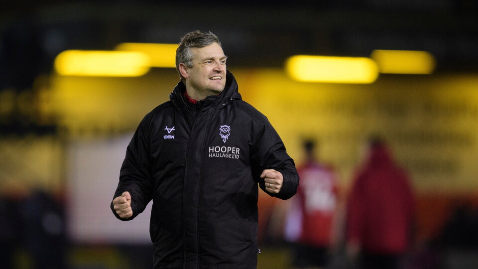 A man in a black jacket stands outside at night, clenching his fists in a celebratory gesture. The background is blurred with warm lights.