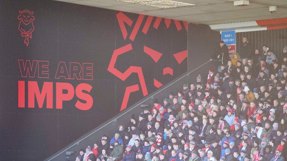 Crowd of football fans in a stadium, clad in winter clothing and red scarves. Bold text and graphic read "We Are Imps" on a black wall. 