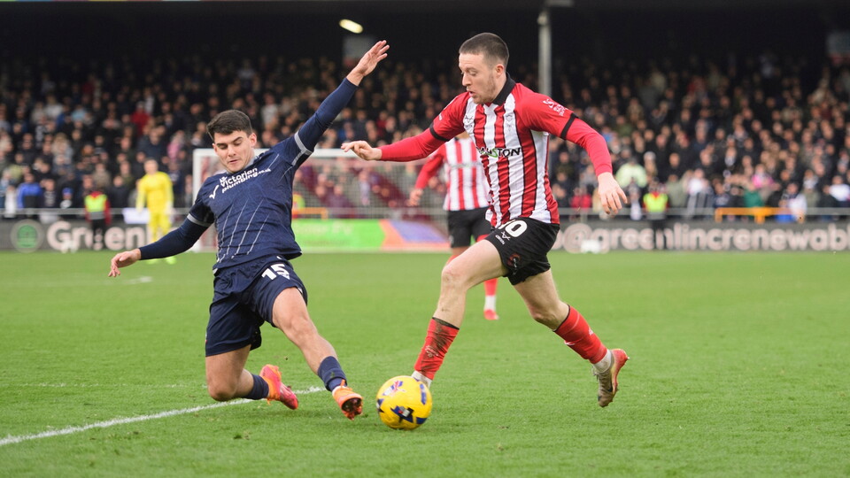 Two players players vie for the ball on a field. The player in red and white stripes dribbles while the player in blue attempts a sliding tackle. 
