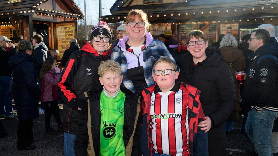 A happy group of fans, wearing sports jerseys and winter jackets, smiles at an outdoor fan village. 