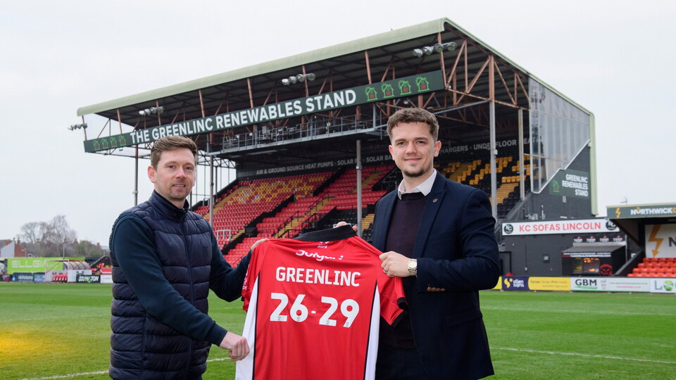 Two men stand on a football field in front of a stadium stand labeled "The GreenLinc Renewables Stand." They're holding a red Lincoln City shirt displaying "GREENLINC 26-29," smiling after announcing a partnership.