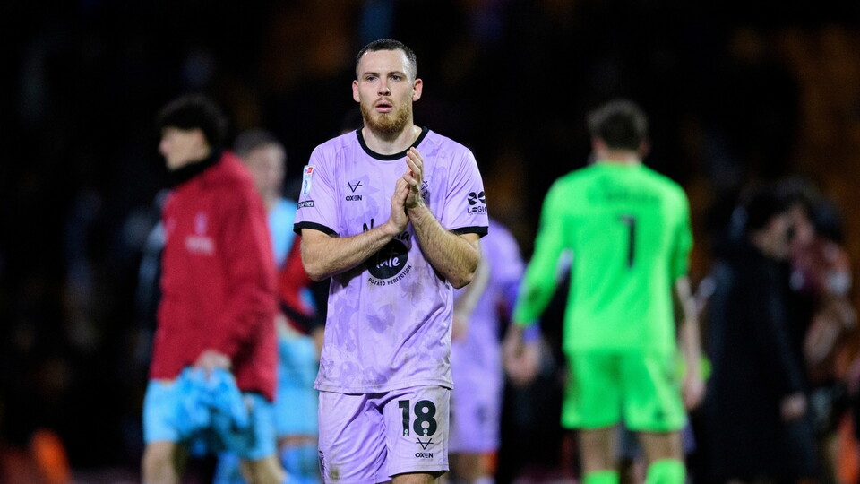 Football player in a violet jersey with number 18 claps hands, looking focused. Teammates and opponents in varied kits are blurred in the background.