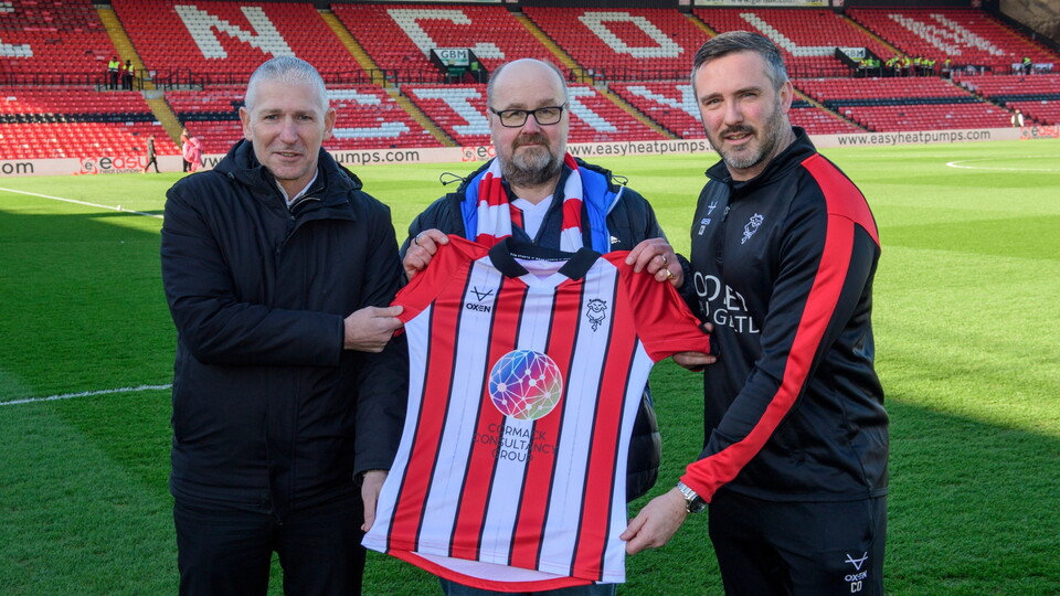 Three men stand on a football pitch, holding a red and white striped Lincoln City shirt, with an empty football stadium in the background. 
