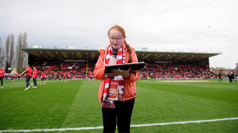 Young person with a scarf and jacket speaks into a microphone on a football pitch, reading from a clipboard. A bustling stadium and spectators are in the background.