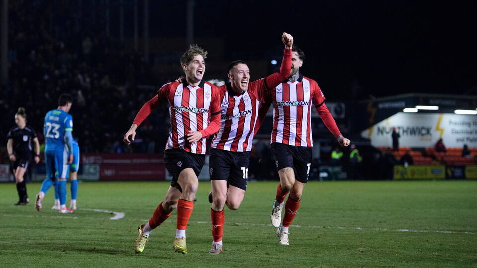 Three football players in red and white striped jerseys celebrate energetically on the pitch at night. A player in blue and a referee are visible in the background.