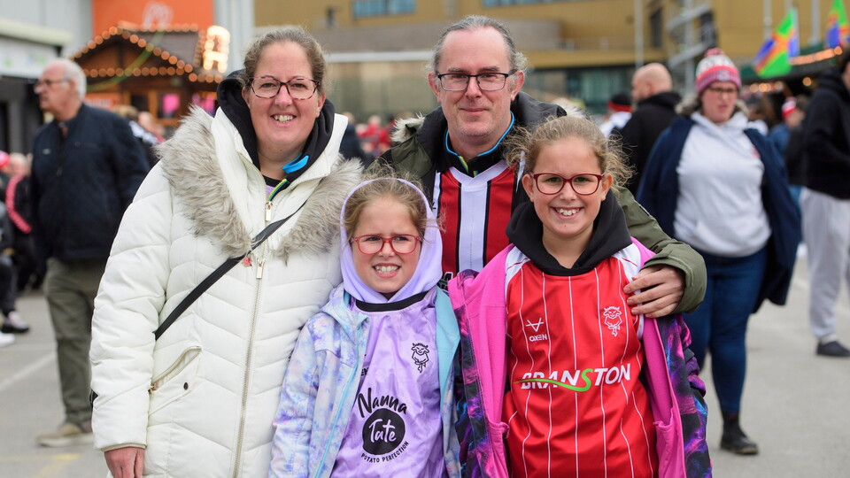 A smiling family of four stands together in a festive outdoor setting. The parents wear jackets, and the children wear sports jerseys. The mood is joyful.