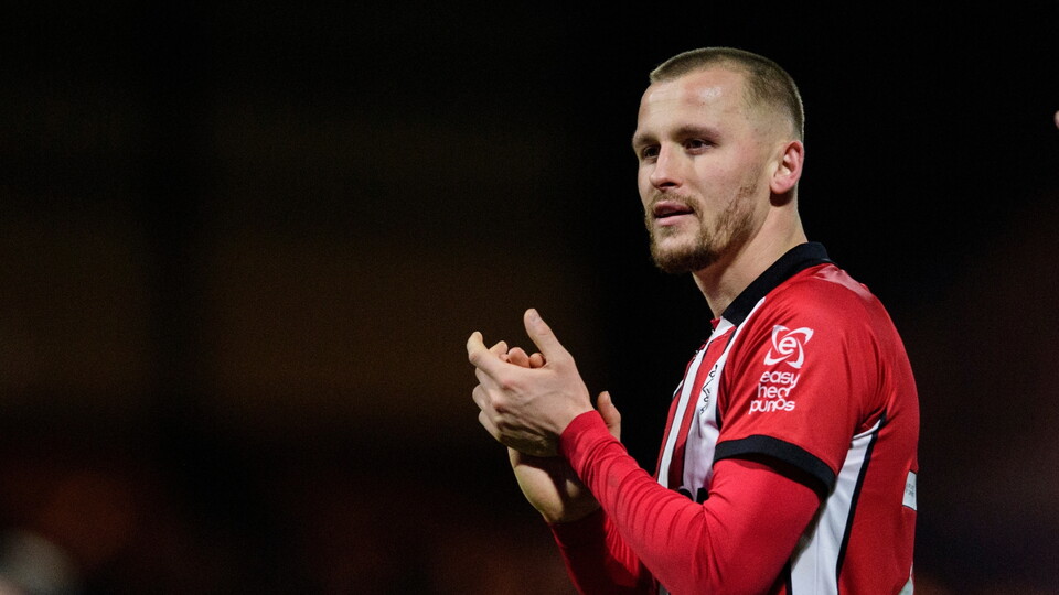 A football player in a red and white striped shirt claps with a focused expression, standing against a dimly lit background.
