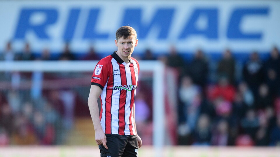 A football player in a red and white striped jersey stands on the pitch, with a blurred crowd and stadium background.
