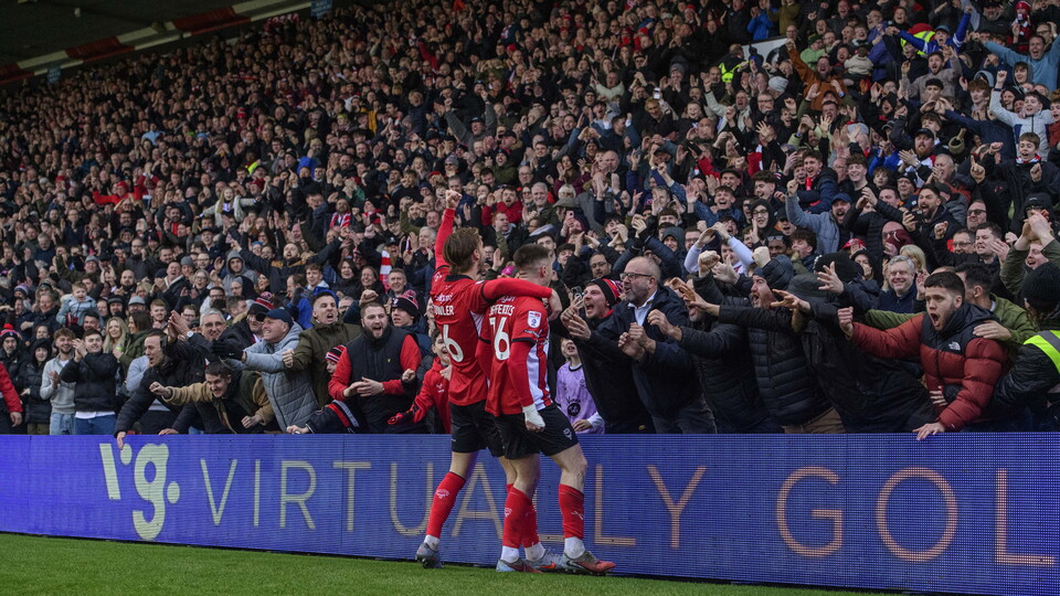Two footballers in red shirts celebrate with a stand full of supporters