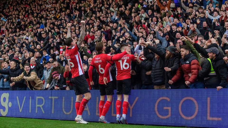 Four players in red jerseys celebrate with an enthusiastic crowd, arms raised in joy, amidst a vibrant stadium atmosphere.