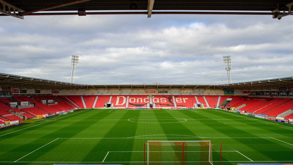 View of Doncaster Rovers' stadium, showing well-maintained green pitch, red seating, and cloudy skies above.