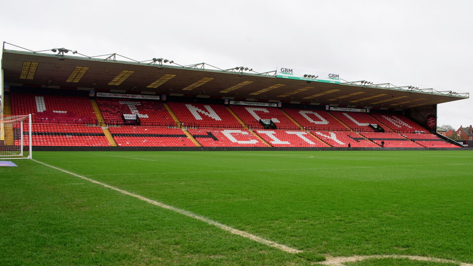 A view of a football stadium with red seating arranged to spell "LINCOLN CITY" on the upper tier and a green pitch in front.