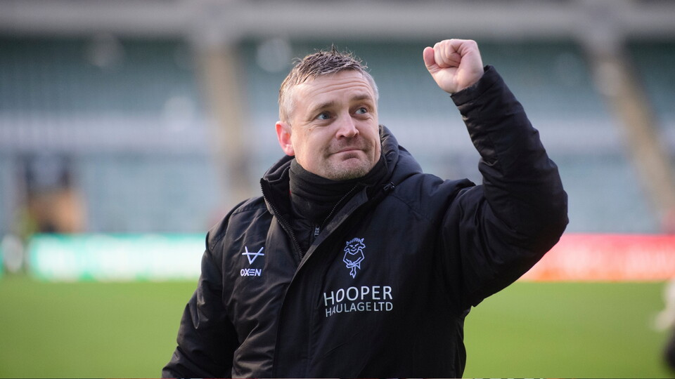 A person in a black jacket raises their fist in celebration at a sports event, with a stadium blurred in the background.