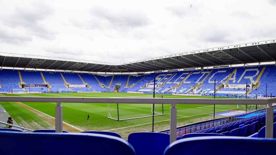 A view of a football stadium with empty blue seats, a green pitch, and goalposts, under a cloudy sky.