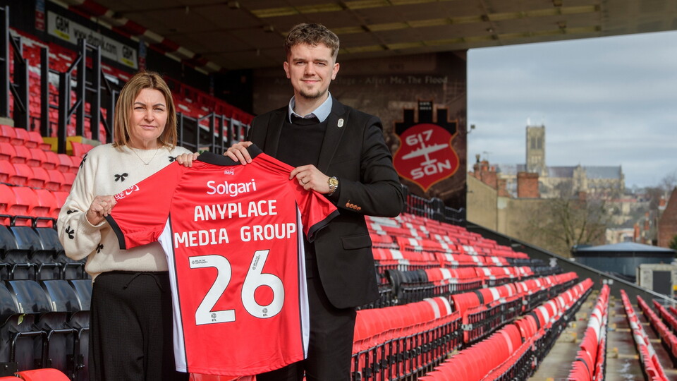 Two people hold up a red football shirt with "ANYPLACE MEDIA GROUP 26" in an empty stadium with red and black seats.