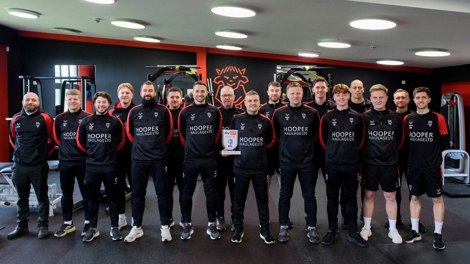 A group of men in black and red training gear standing in a gym, one holding a Sky Bet League One Manager of the Month trophy.