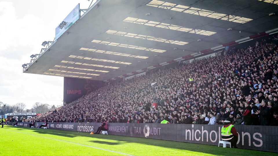 Crowd of spectators fills the covered stadium stand at a daytime football match.