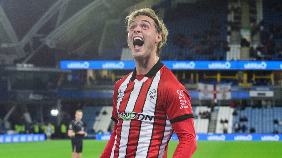 Football player in red and white striped jersey celebrating with clenched fists on stadium pitch during nighttime match.