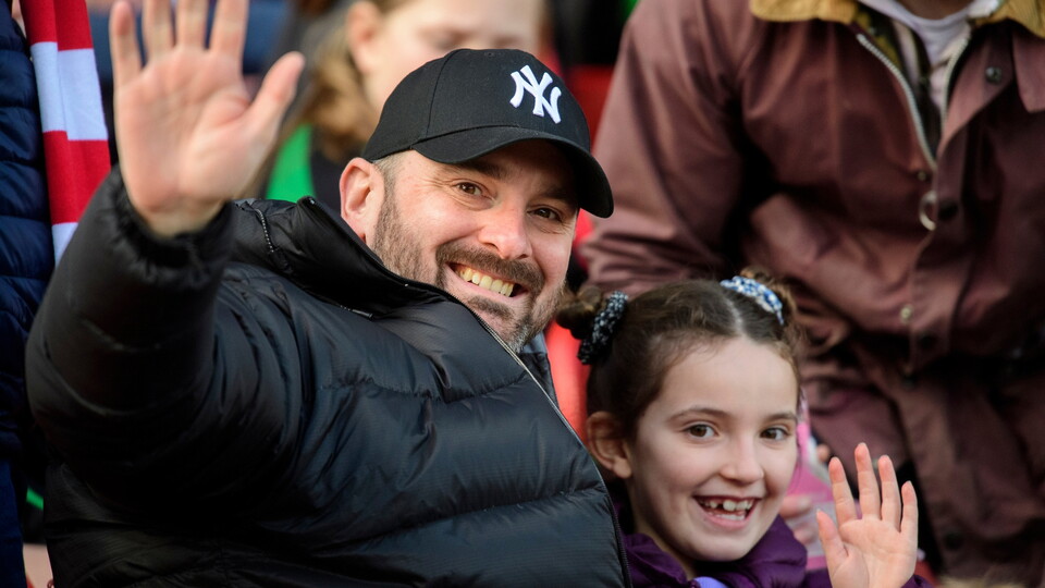 Two people waving hands, wearing winter jackets, in a crowded outdoor setting.