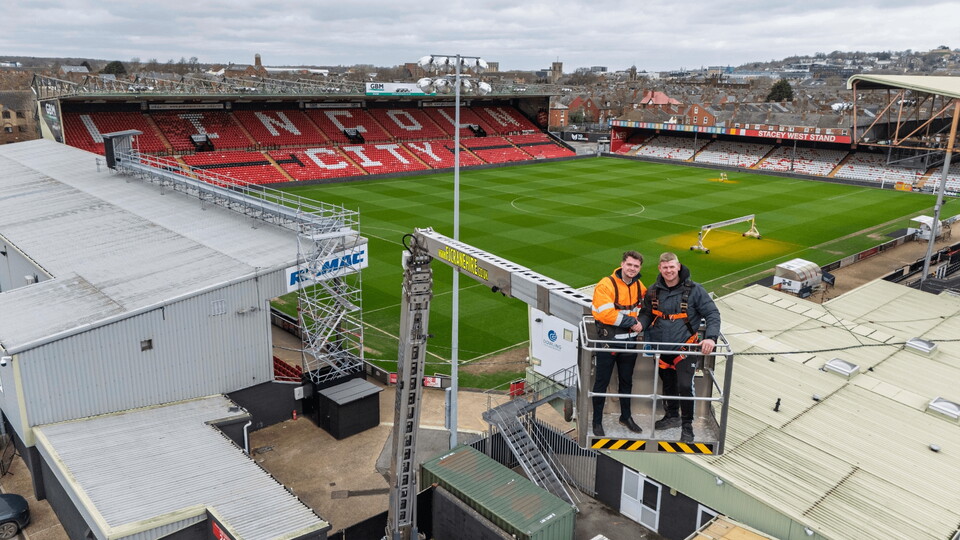 Two men in safety gear on a crane overlooking an empty stadium.