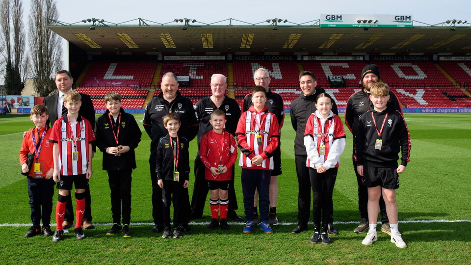 A group of children and adults posing on a football pitch with ‘Lincoln’ visible on the red stadium seats in the background. 