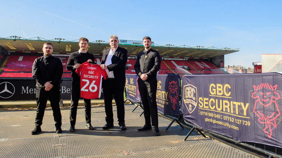 Four men stand inside a football stadium, holding a red jersey with 'GBC SECURITY' and the number 26. Bright, sunny day; security banners are visible.