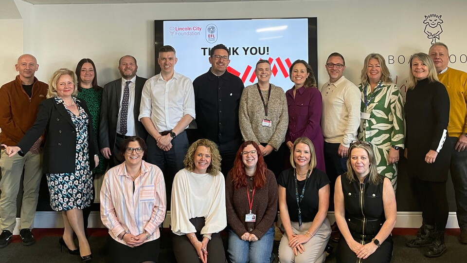 A group of people, smiling, pose in two rows in front of a screen displaying "Thank You!". They are in a cheerful and professional setting.