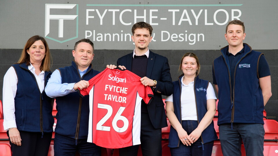 Five people stand in a stadium holding a red football shirt that says 'Fytche Taylor 26.' They are smiling under a 'Fytche-Taylor' sign.