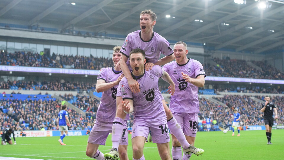 Football players in violet kits joyfully celebrate a goal on a vibrant green pitch in a packed stadium.
