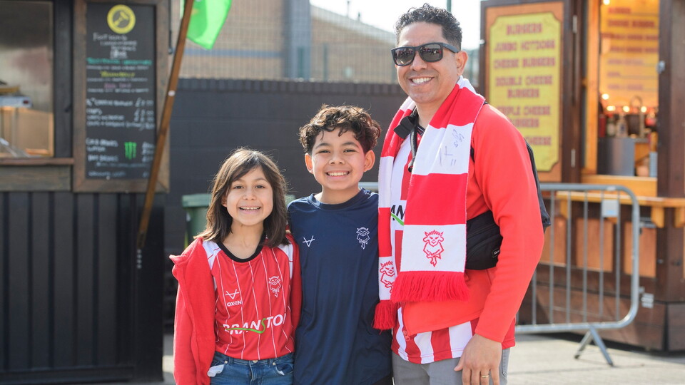 A smiling man and two children wear matching red football shirts and scarves. They stand outside a food stall.