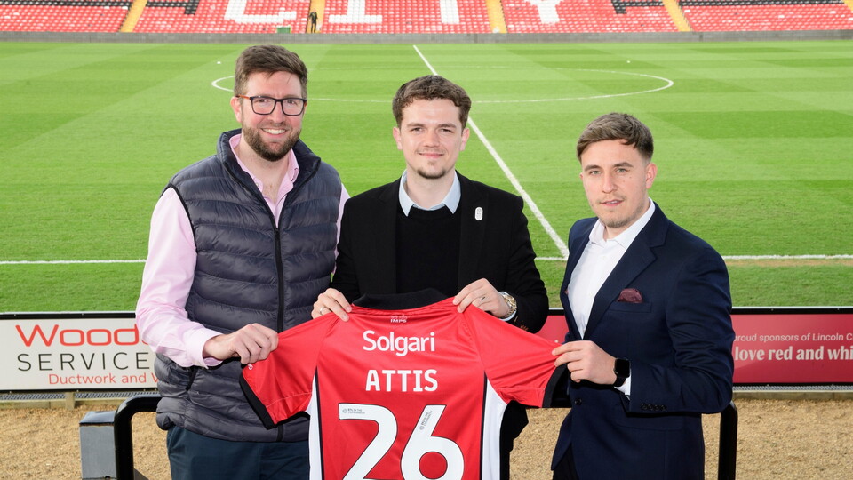 Three men smiling and holding a red football shirt with "ATTIS 26" on the back. They're standing in a stadium with green pitch and red seats in the background.
