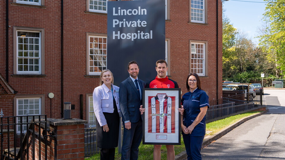Four people stand smiling in front of Lincoln Private Hospital, holding a framed red and white striped sports jersey. The atmosphere is celebratory.