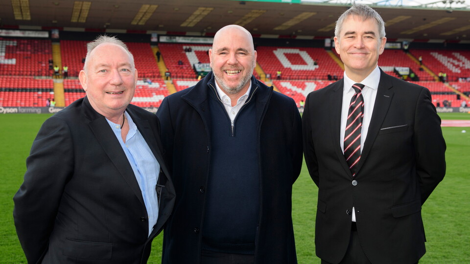 Three men in suits pose in front of a football pitch