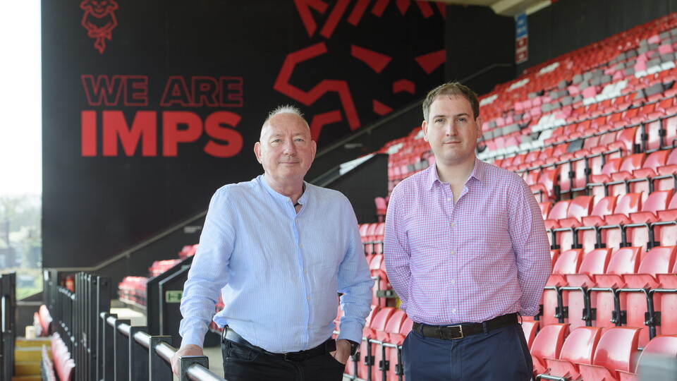 Two men pose in front of a sign which reads We Are Imps