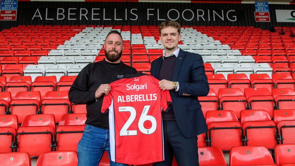 Two men stand in a stadium holding a red football shirt with "Alberellis" and "26" on it. The seating is red and white.
