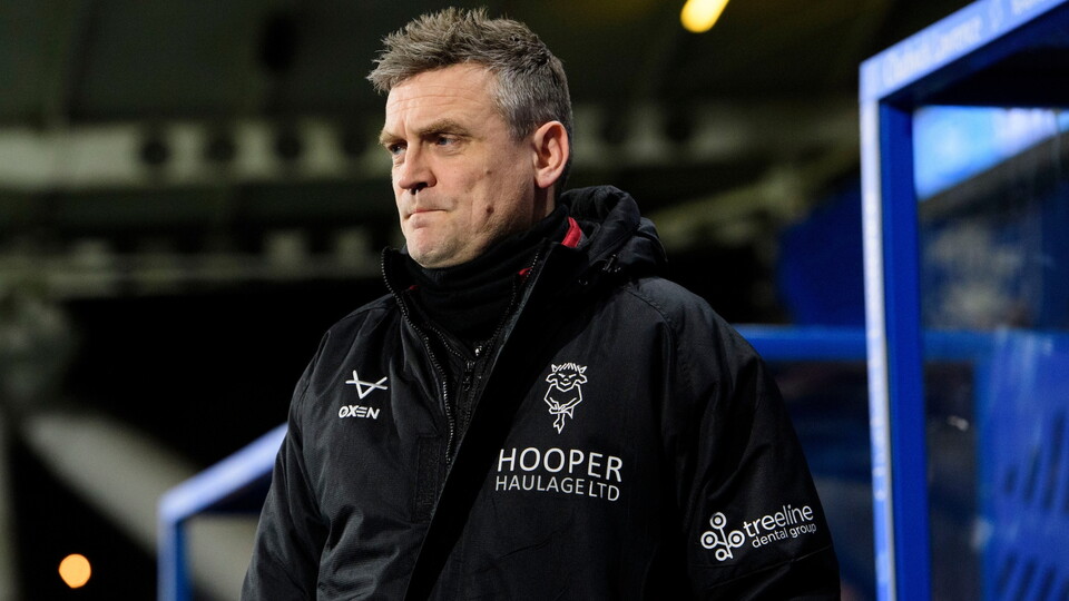 A man wearing a black jacket with logos, stands pensively in a sports stadium. The blue seating area and stadium roof are visible in the background.