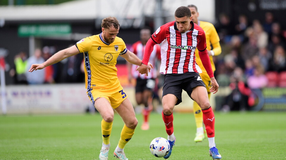 Two football players compete for the ball on a grassy field. One wears a yellow kit, the other a red and white striped jersey.