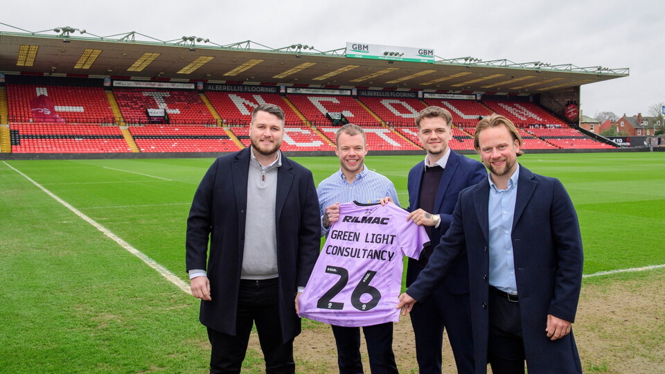 Four smiling men in coats stand on a football pitch, holding a violet jersey with the number 26. The background shows empty red stadium seats.