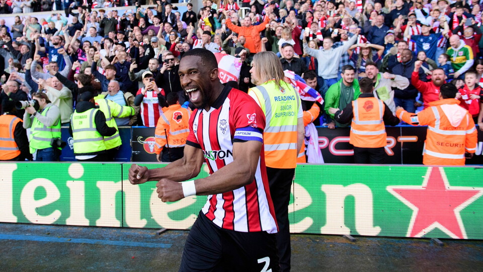 A football player in a red and white shirt celebrates energetically with clenched fists. The cheering crowd and security in high-visibility vests are visible behind him.