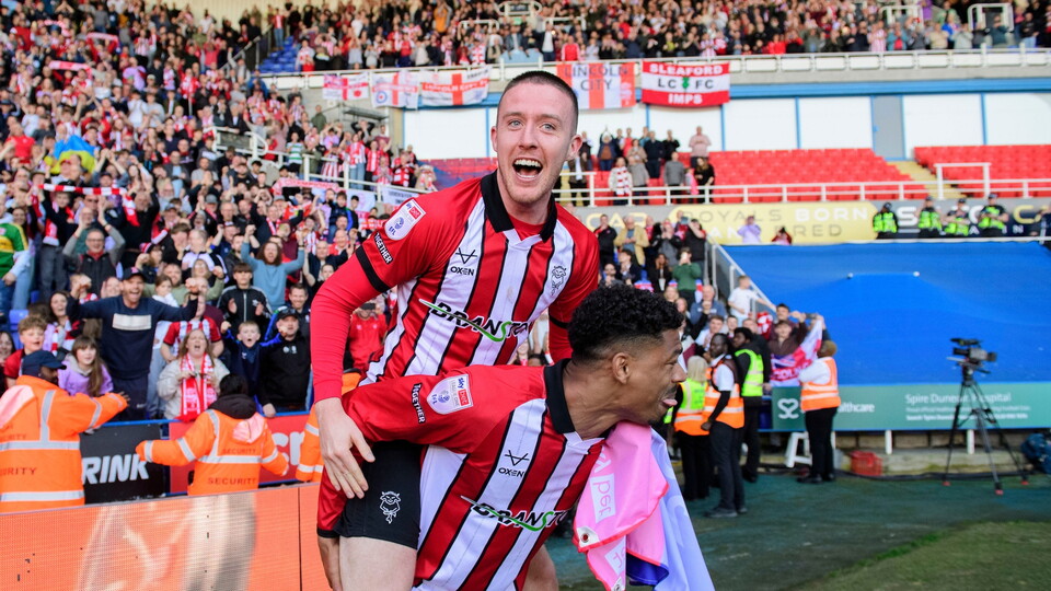 Two football players in red and white shirts celebrate, with one jumping on the other's back. A joyful crowd and security staff are in the background.