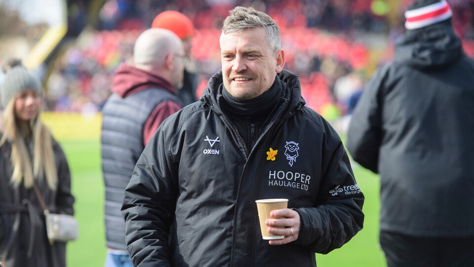 Man in a black jacket with logos holds a coffee cup on a football pitch. Background shows blurred spectators and a person in a beanie hat. 