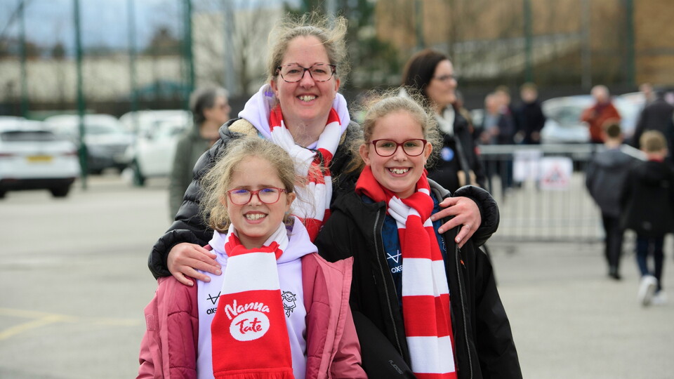 A smiling woman and two girls, all wearing red and white scarves, stand outdoors.