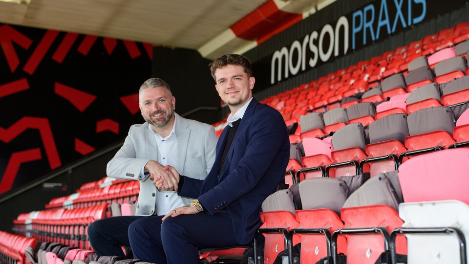 Two men in suits shake hands while sitting in a stadium with red and black seats. 